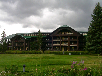 [A four story brown building with a green metal roof. There are two sections which stick out from the main building which appear to be rooms with balconies. There is a lush green lawn in front with some wildflowers growing at the very front of the photo (near the road).]